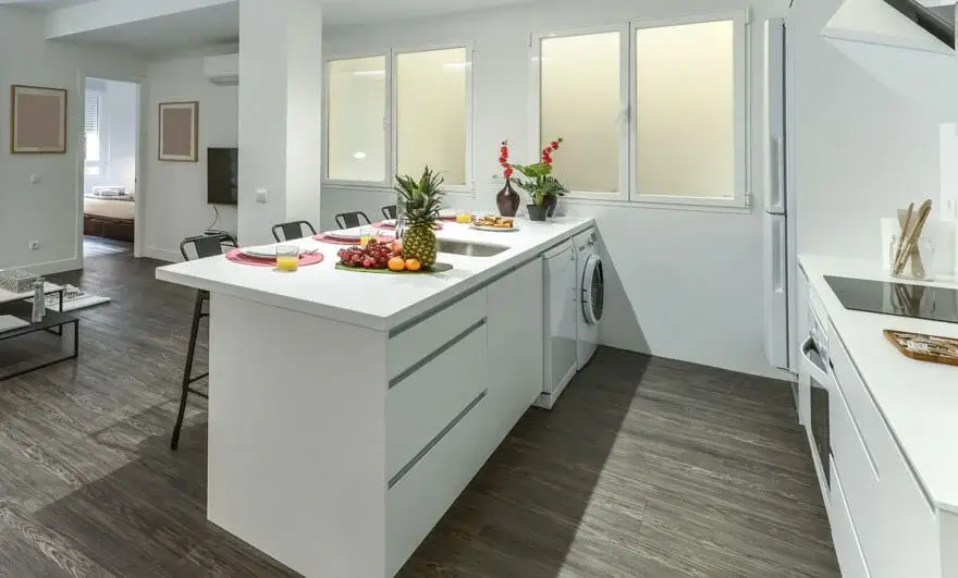 Parallel modular kitchen design featuring a kitchen island with a white countertop, cabinets with drawers, black stools, and a breakfast setup with assorted fruits and glasses of orange juice.